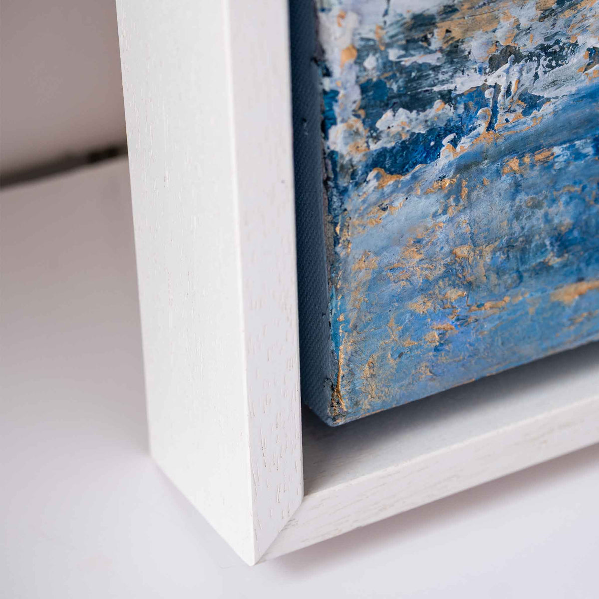 Close-up of a white wooden picture frame corner holding a textured blue and gold abstract canvas painting.