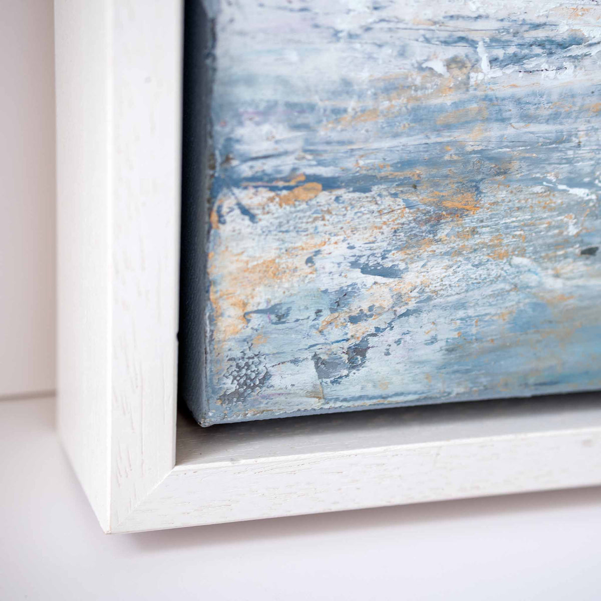 Close-up of a framed abstract canvas print showing the corner of the white frame and the textured painting with blue, gray, and gold tones.