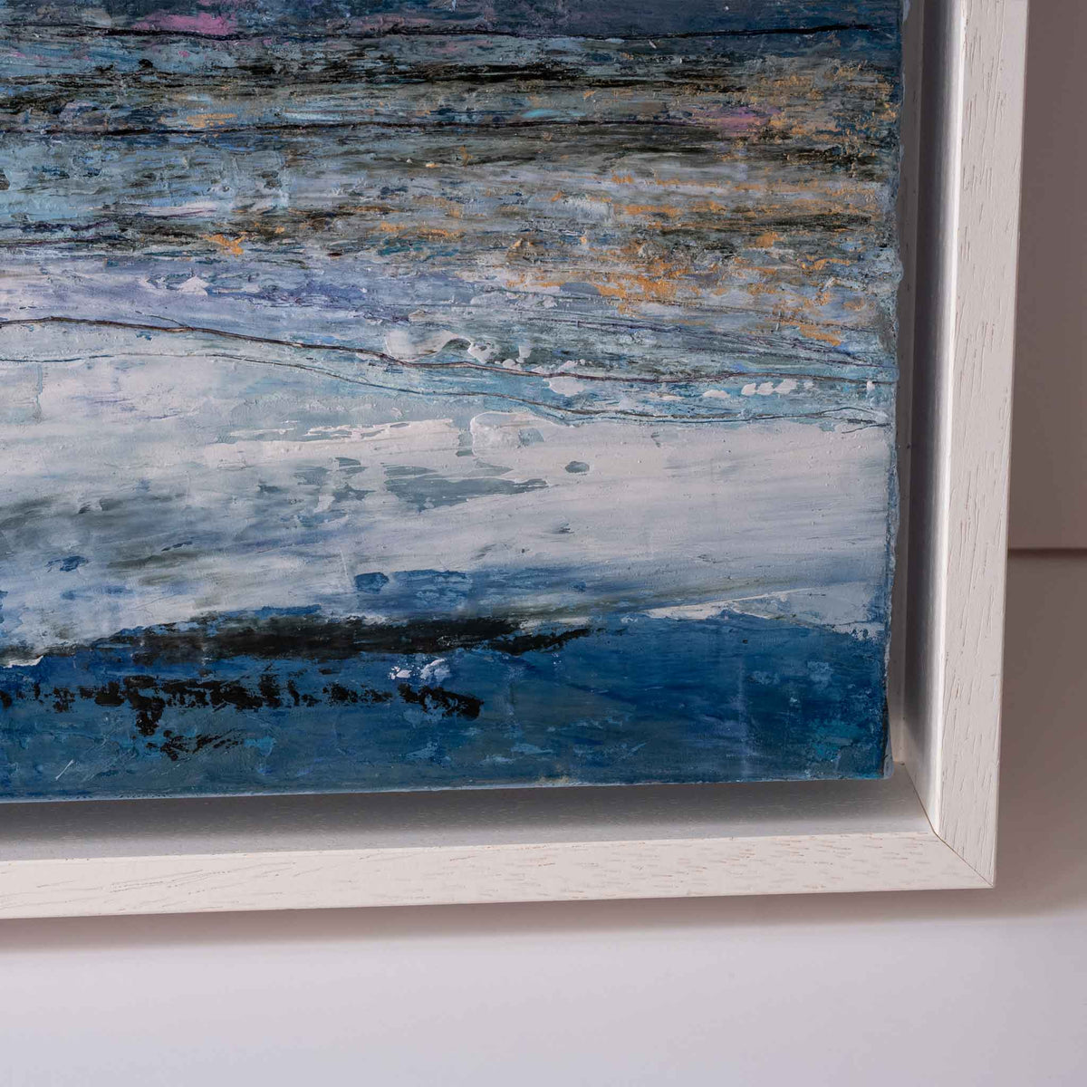 Close-up of a framed abstract painting with layered blue, white, gray, and black brushstrokes, set in a white wooden frame against a light wall.