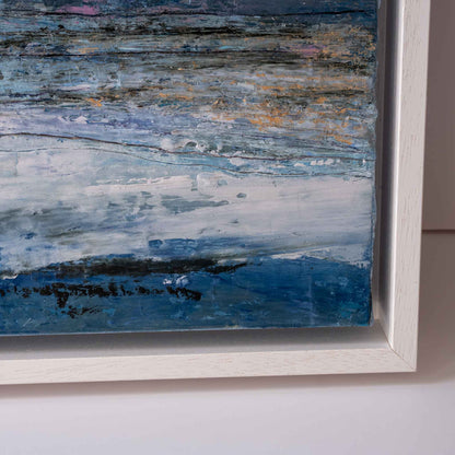 Close-up of a framed abstract painting with layered blue, white, gray, and black brushstrokes, set in a white wooden frame against a light wall.