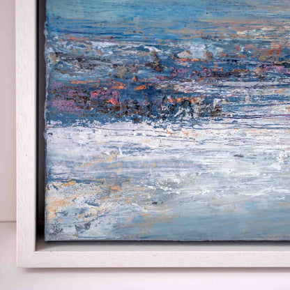Close-up of a framed abstract painting with textured brushstrokes in blue, white, gray, and touches of pink, purple, and orange, shown in a white frame against a light wall.